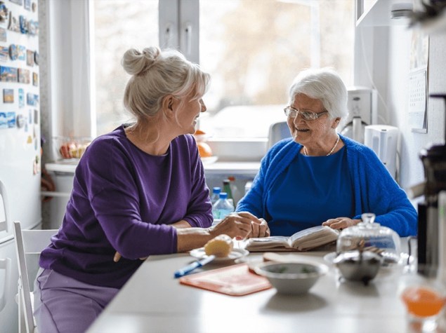 Mujer adulta acompaña a mujer mayor en la cocina, conversando y revisando un cuaderno