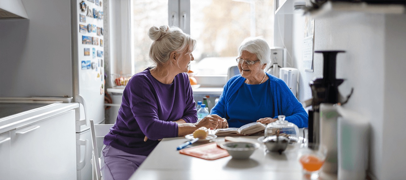 Mujer adulta acompaña a mujer mayor en la cocina, conversando y revisando un cuaderno