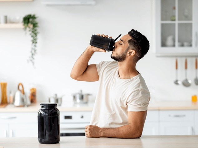 Hombre bebe batido de proteínas con vaso mezclador, junto a bote de suplemento en cocina.