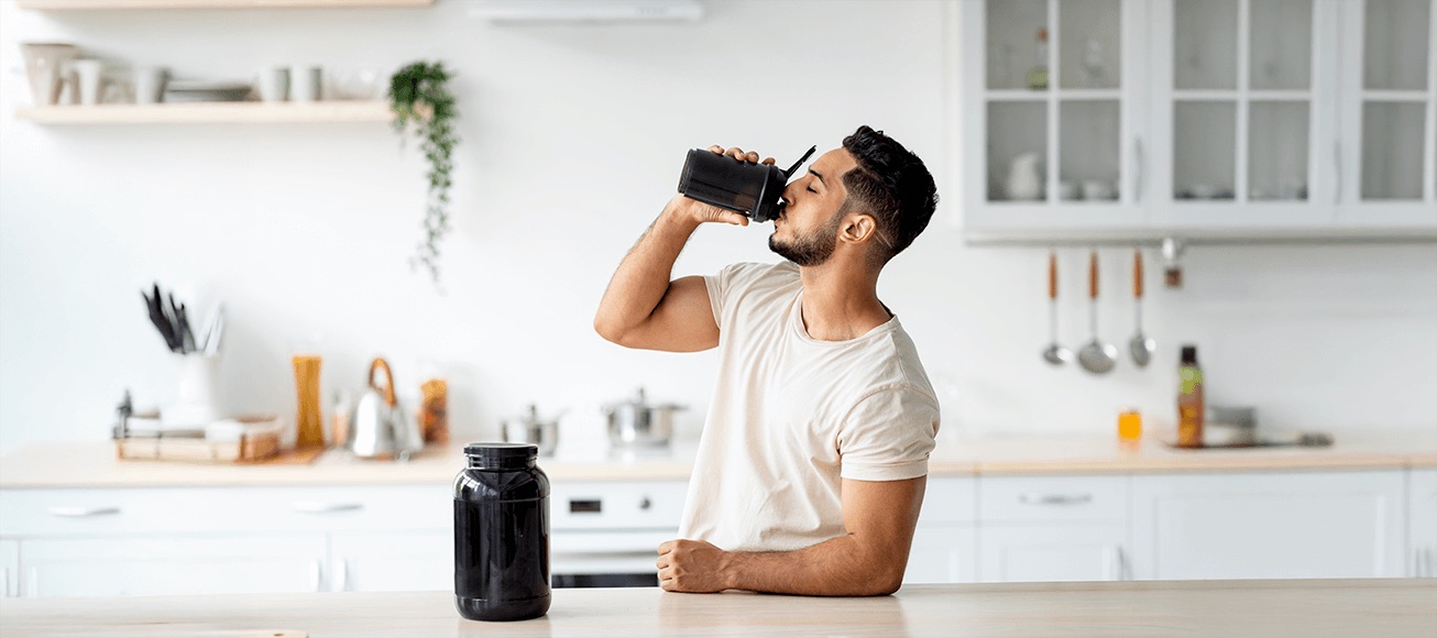 Hombre bebe batido de proteínas con vaso mezclador, junto a bote de suplemento en cocina.
