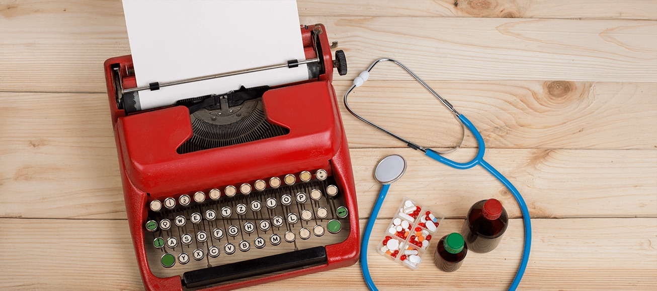 Máquina de escribir roja con papel, estetoscopio azul, pastillas y frascos sobre mesa de madera.