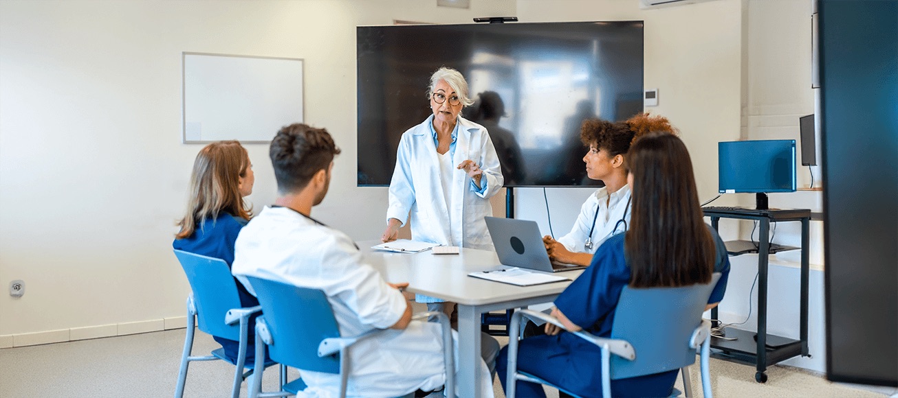 Mentora médica guía a estudiantes de medicina en aula clínica, con pantalla y portátil.