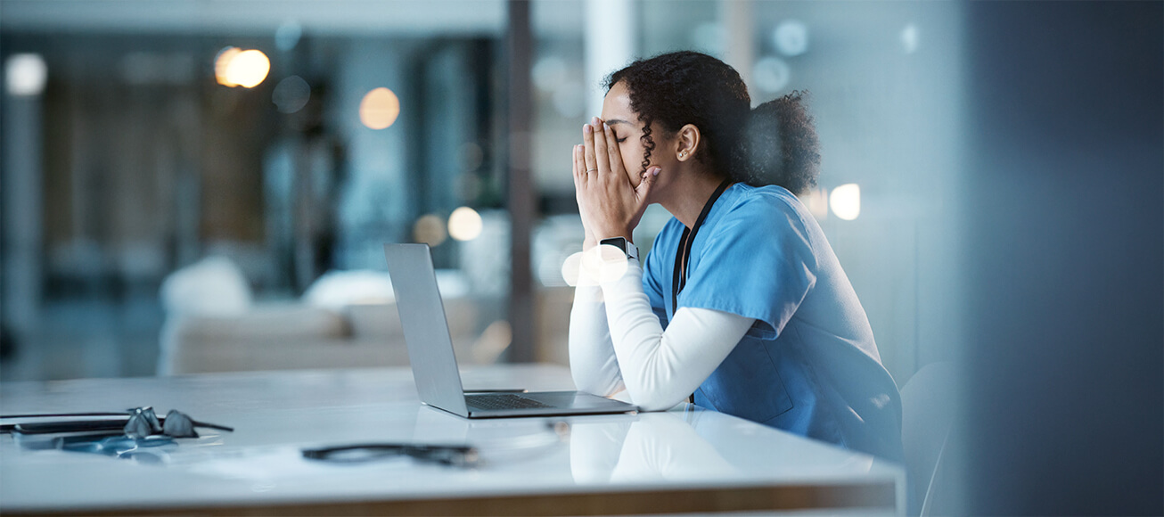 Profesional sanitario con pijama medico, agotado frente a portatil en hospital, manos cubriendo rostro, guardia nocturna.