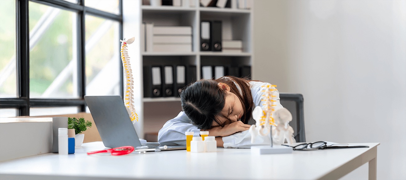 Profesional de salud dormida en escritorio con laptop y modelo de columna