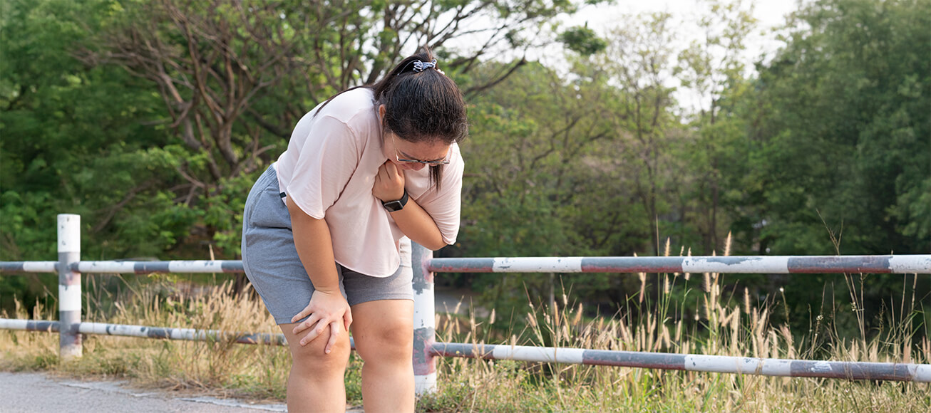 Mujer joven en parque se detiene durante ejercicio, con dolor torácico y dificultad para respirar.
